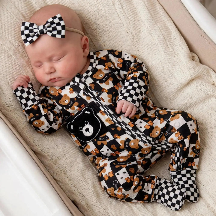 Baby wearing a black and white checkered convertible romper with teddy bear pattern, sleeping on a beige blanket.