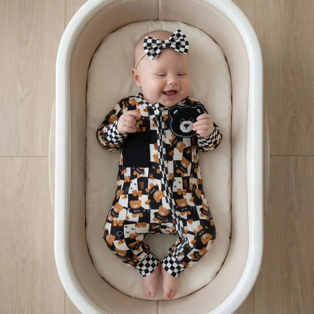 Baby in a sensory convertible  onesie and bow tie laying in a crib on a wooden floor. Looking at a teddy bear patch