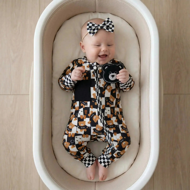 Baby in a sensory convertible  onesie and bow tie laying in a crib on a wooden floor. Looking at a teddy bear patch